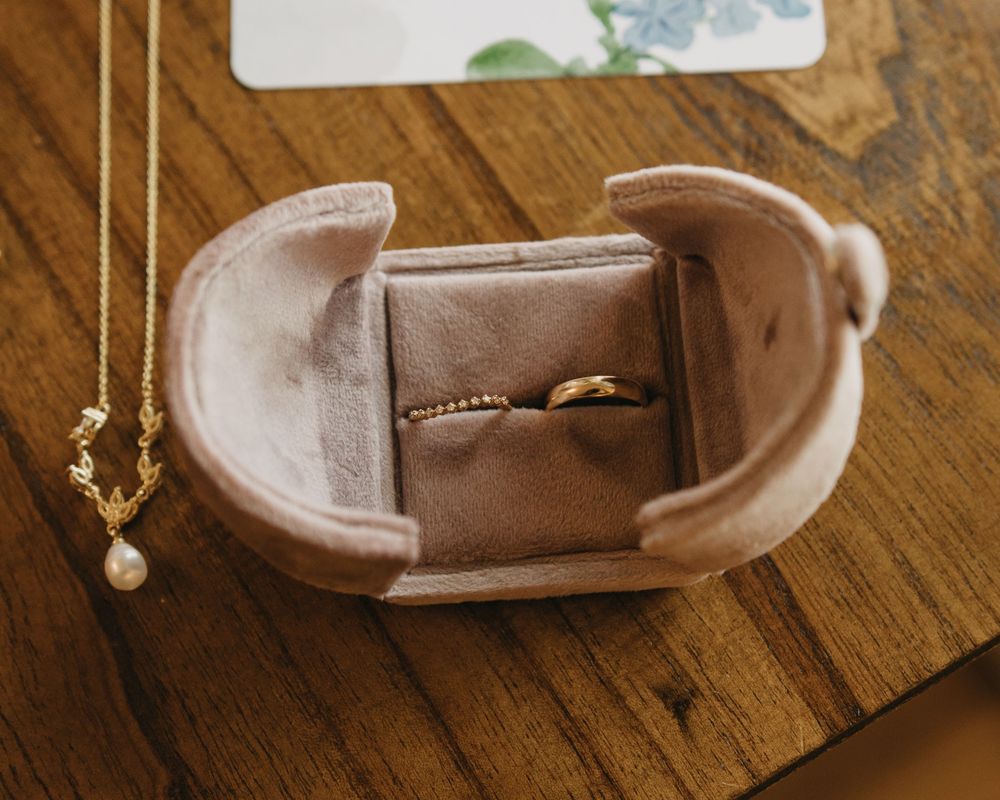 Two gold wedding bands in a velvet ring box beside a delicate pearl pendant necklace on a wooden table.