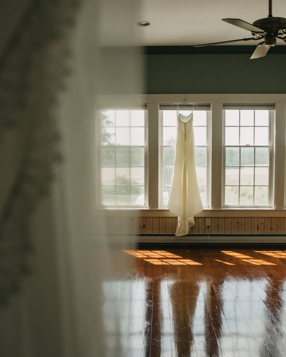 Elegant white wedding dress hanging in sunlit room with hardwood floors.