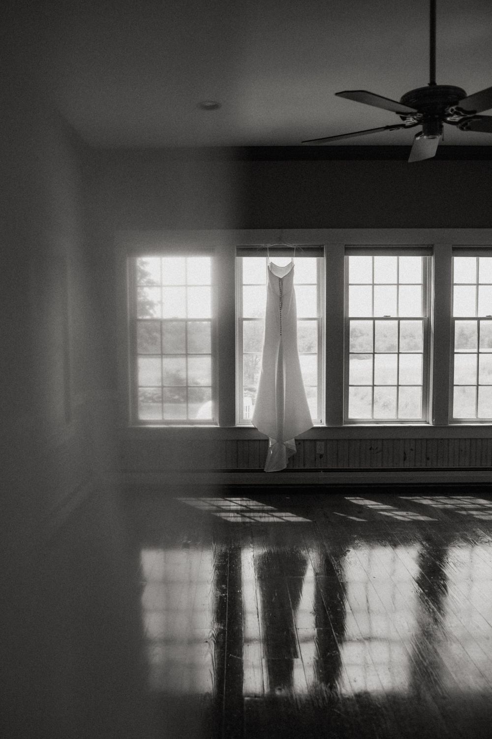 Black and white photo of a wedding dress hanging in a sunlit window with hardwood floors.