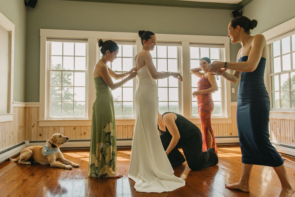 Bridesmaids in colorful gowns help bride with final dress adjustments in sunlit room with a dog nearby.