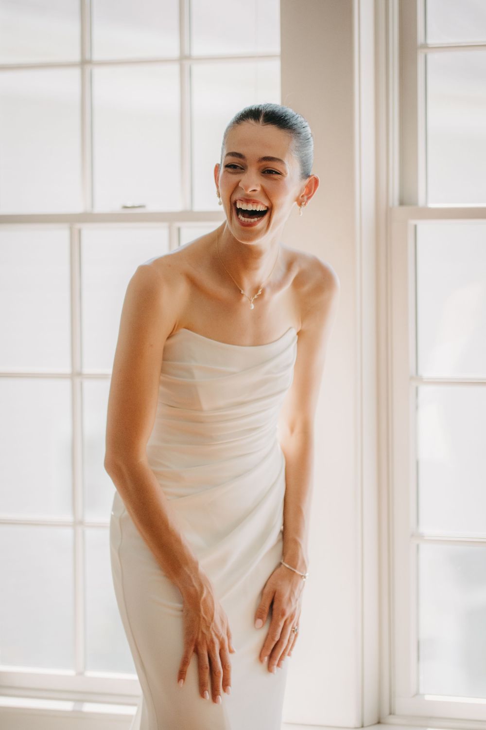 Radiant bride in strapless white wedding dress laughing joyfully while standing before bright windows.