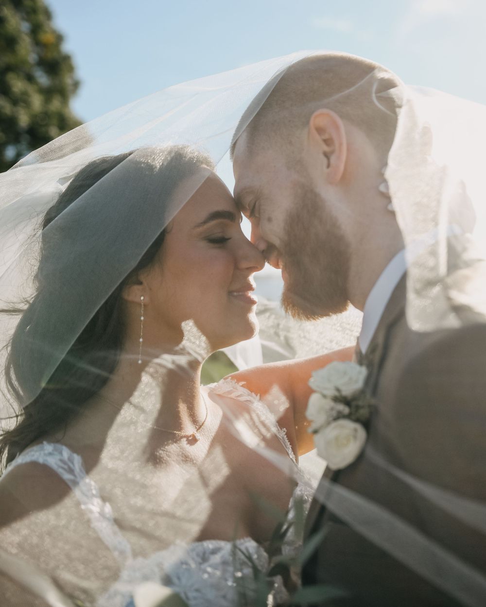 Bride and groom touching foreheads tenderly under a flowing veil in soft golden sunlight.