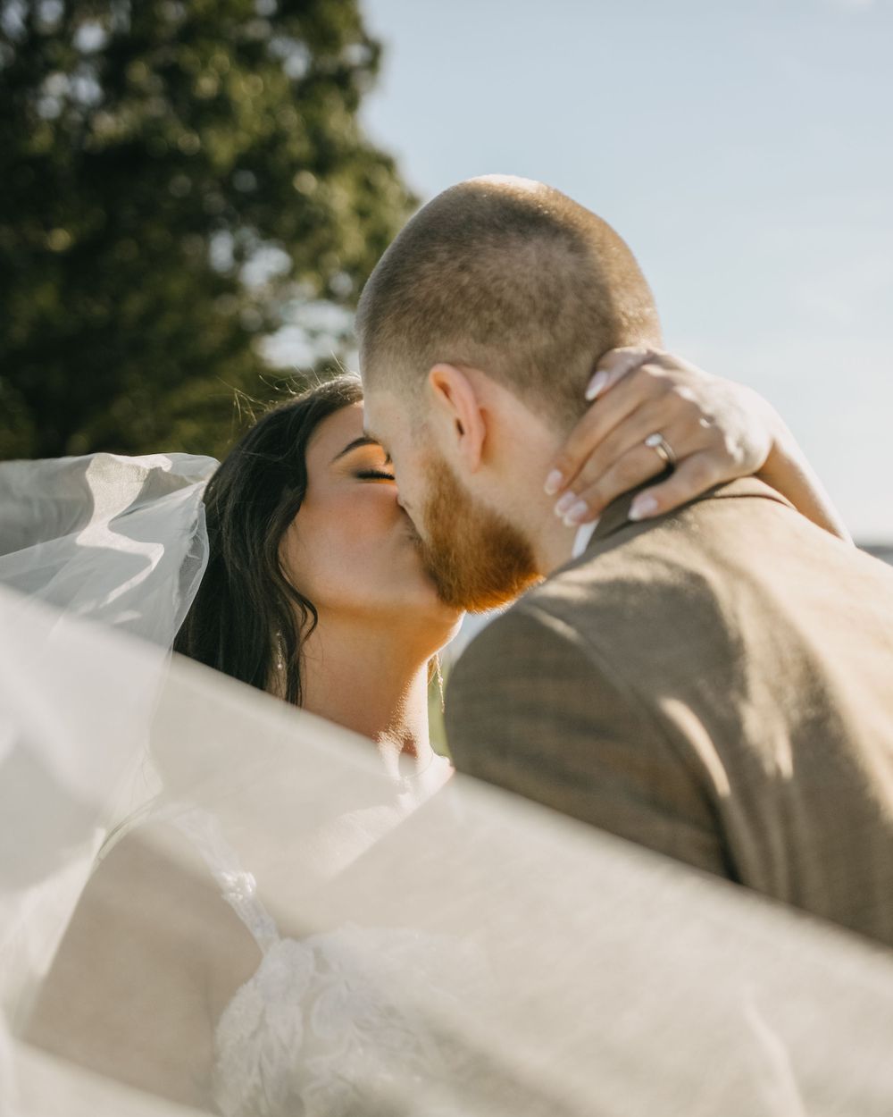 Bride and groom sharing a romantic kiss outdoors with wedding veil flowing in the sunlight.