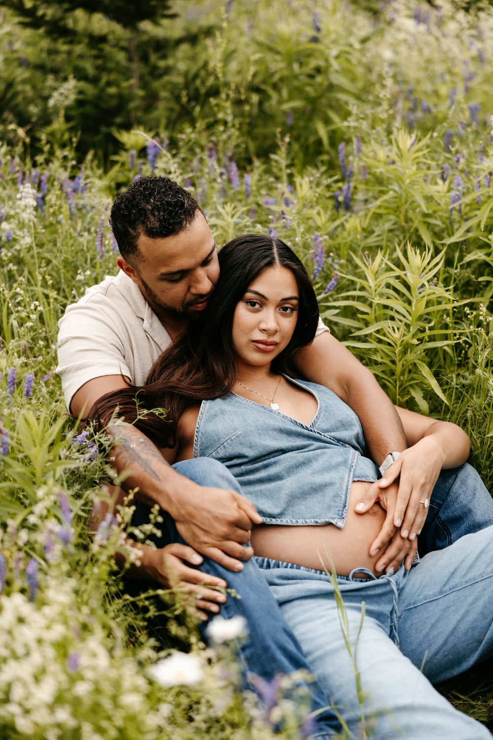 A couple sits in a wildflower field, man embracing pregnant woman in denim outfit, maternity photo session outdoors.