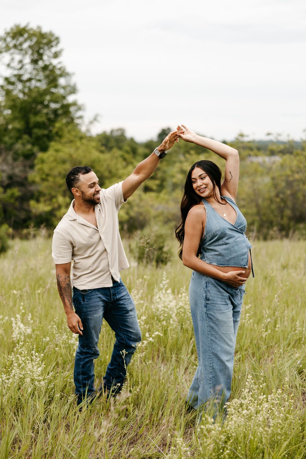 A couple dances in a lush green field, the pregnant woman in denim smiling as her partner spins her around.