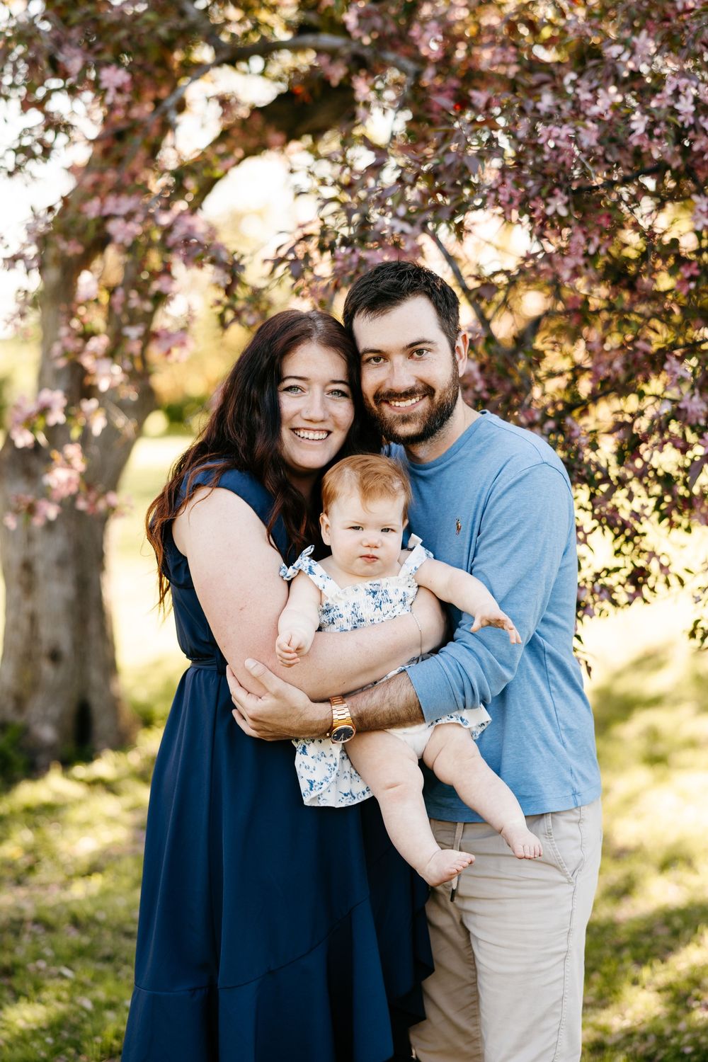 Smiling family with baby posed under pink flowering tree outdoors in spring.