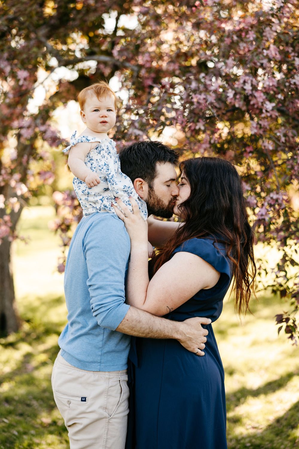 A couple kisses while holding their baby girl under blooming pink cherry blossom trees in a park.