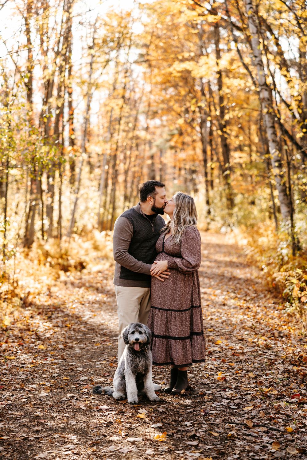 A couple embraces on an autumn forest path with their gray dog standing beside them on fallen leaves.
