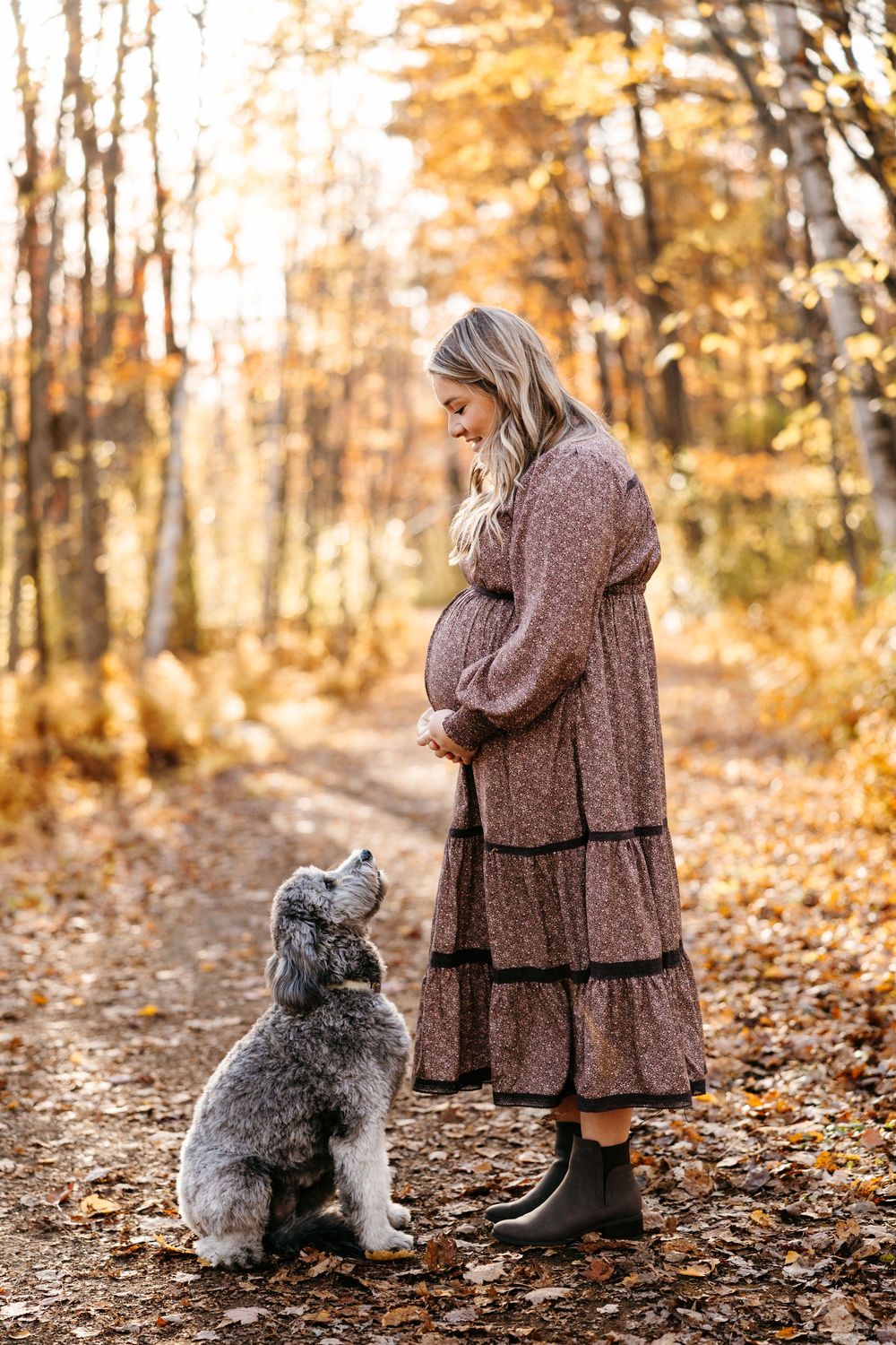 A small grey dog sits beside someone wearing a brown coat on a leaf-covered autumn forest path.