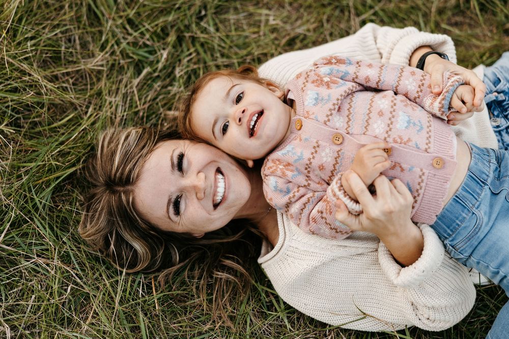 A person lying in green grass wearing a pink sweater and denim jeans while smiling and laughing.