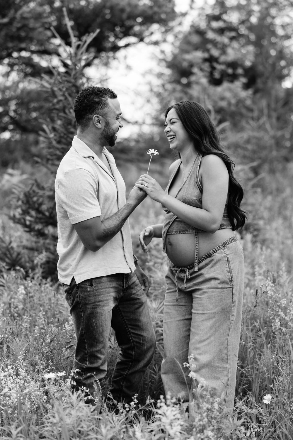 Black and white photo of a happy pregnant couple sharing a flower in a lush outdoor field.