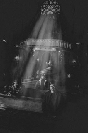 Black and white photo of a band posing inside a dark cathedral with dramatic light rays.