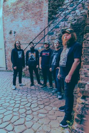 Five heavy metal band members pose in band tees against a rustic brick wall and cobblestone alley.