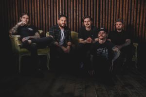 Five rock band members pose together on vintage green chairs against a dark wooden backdrop.