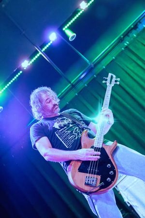 A bassist plays electric bass guitar under vibrant blue and green stage lights during a live concert.