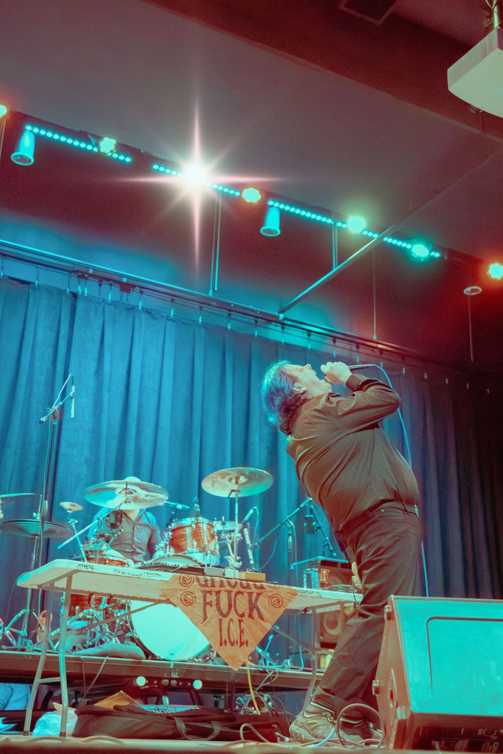 A vocalist performs passionately on stage under teal and red lights with a drum kit visible behind.
