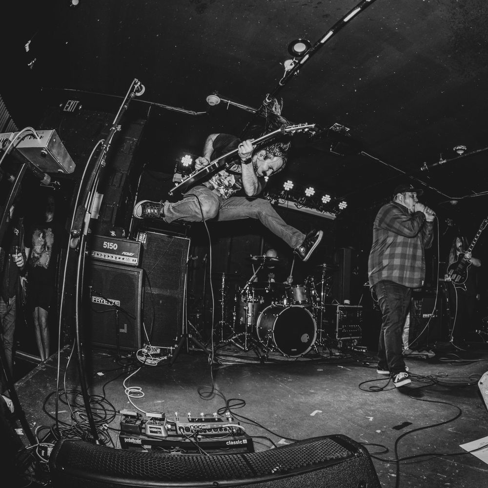 Black and white live concert photo of guitarist jumping on stage with band performing under bright lights.