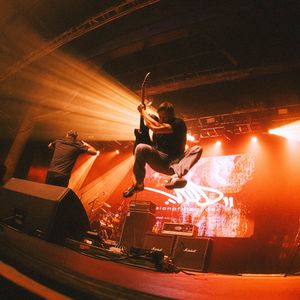 Guitarist leaps mid-air on a brightly lit orange stage during an energetic live rock concert.