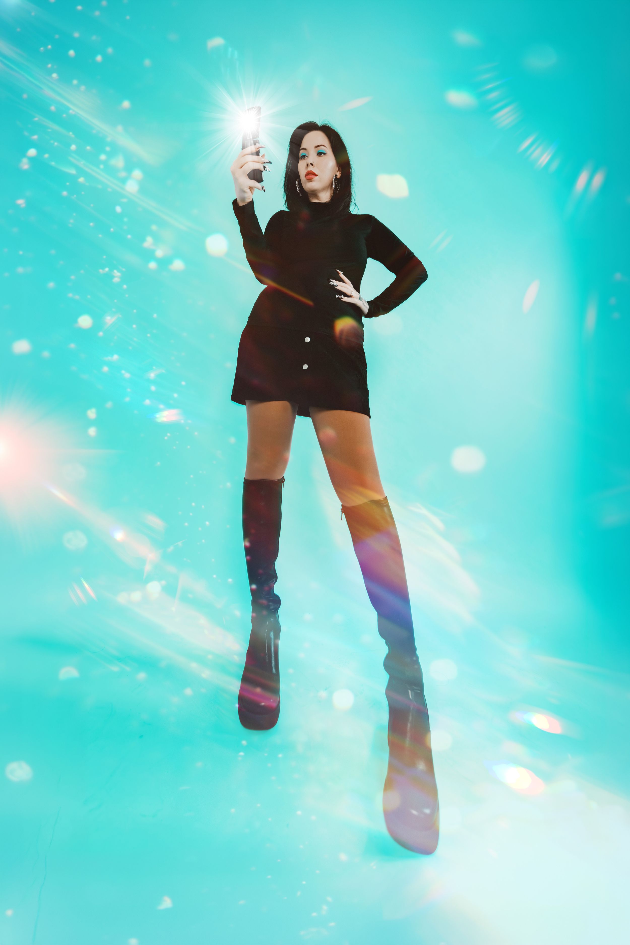 Woman in black mini dress and thigh-high boots holding glowing light on vibrant teal background with bokeh effects.