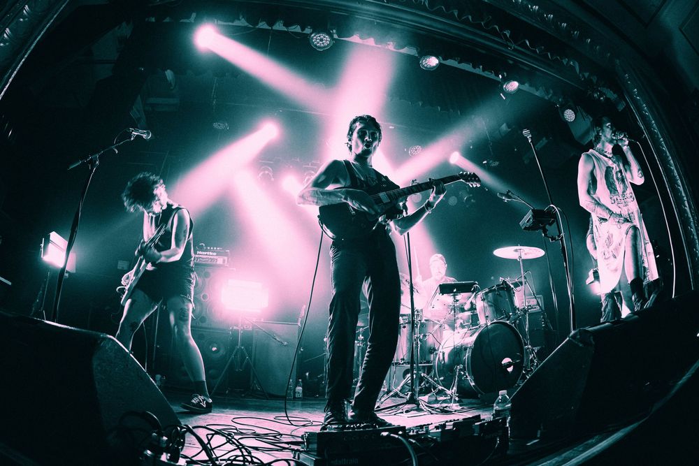 Rock band performs on stage under pink and blue lights captured with fisheye lens at live concert.