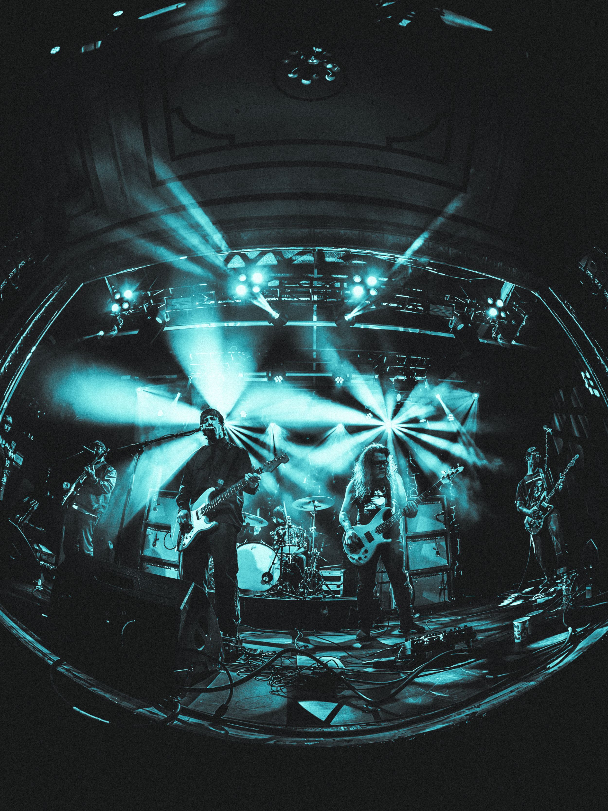 Wide-angle fisheye view of rock band performing on stage with dramatic teal lighting and light beams overhead.