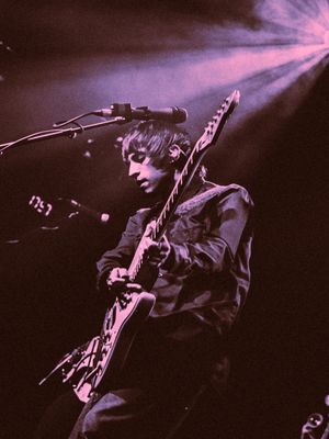 Guitarist performing on stage bathed in purple and pink spotlight rays during a live concert performance.