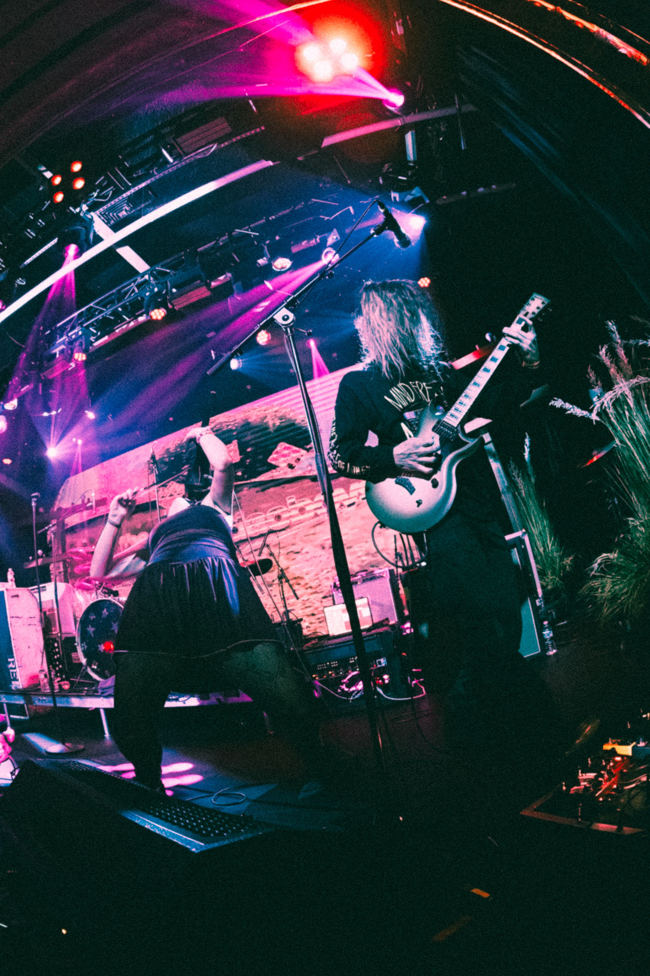Guitarist with long hair performs on stage under vibrant pink and purple lights at a live rock concert.