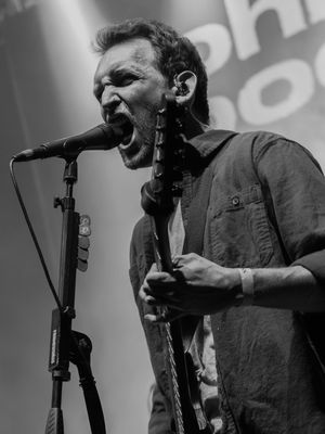 Black and white photo of a bearded male musician singing passionately into a microphone while playing guitar on stage during a live concert.