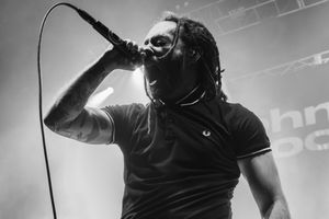 Black and white photo of a dreadlocked male vocalist singing into a microphone on stage during a live concert.