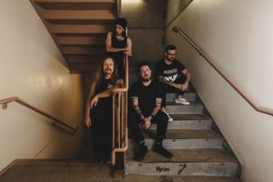 Three band members pose on concrete stairs in a dimly lit stairwell, dressed in black clothing.