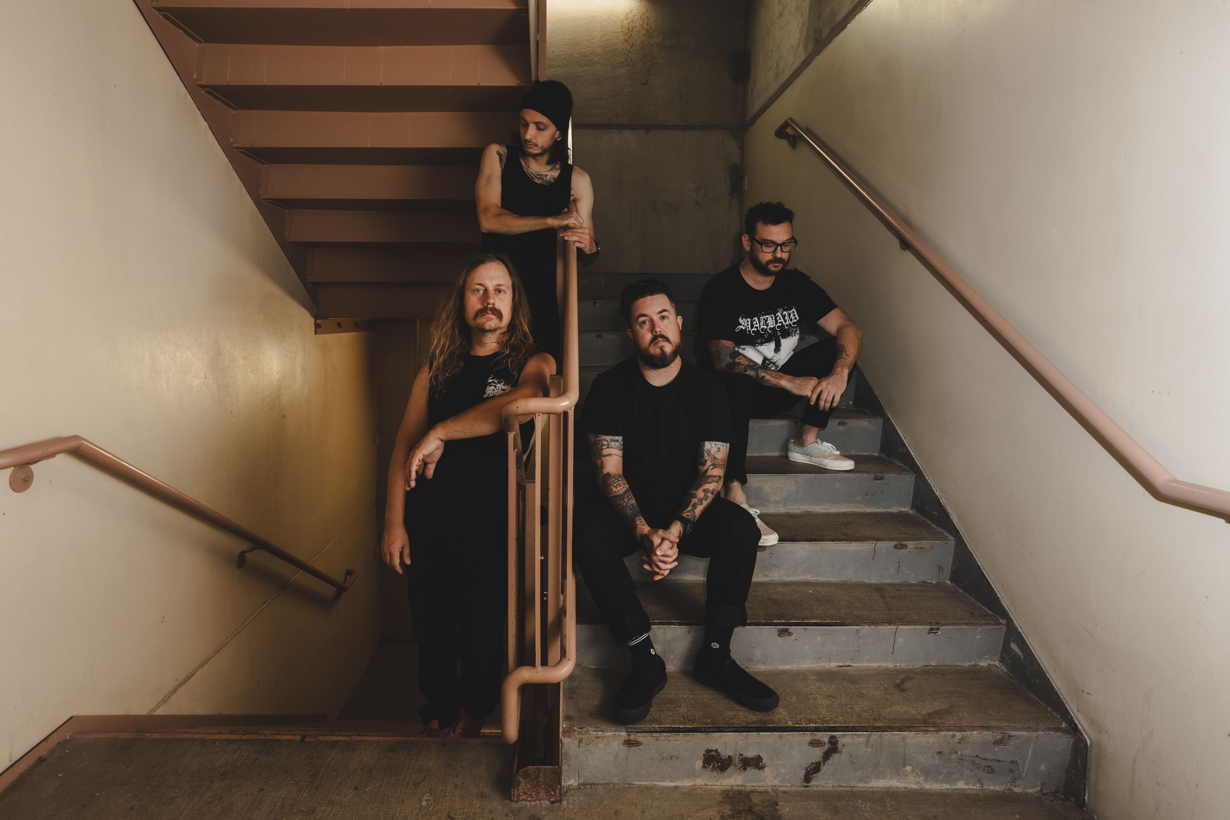 Three band members pose on concrete stairs in a dimly lit stairwell, dressed in black clothing.