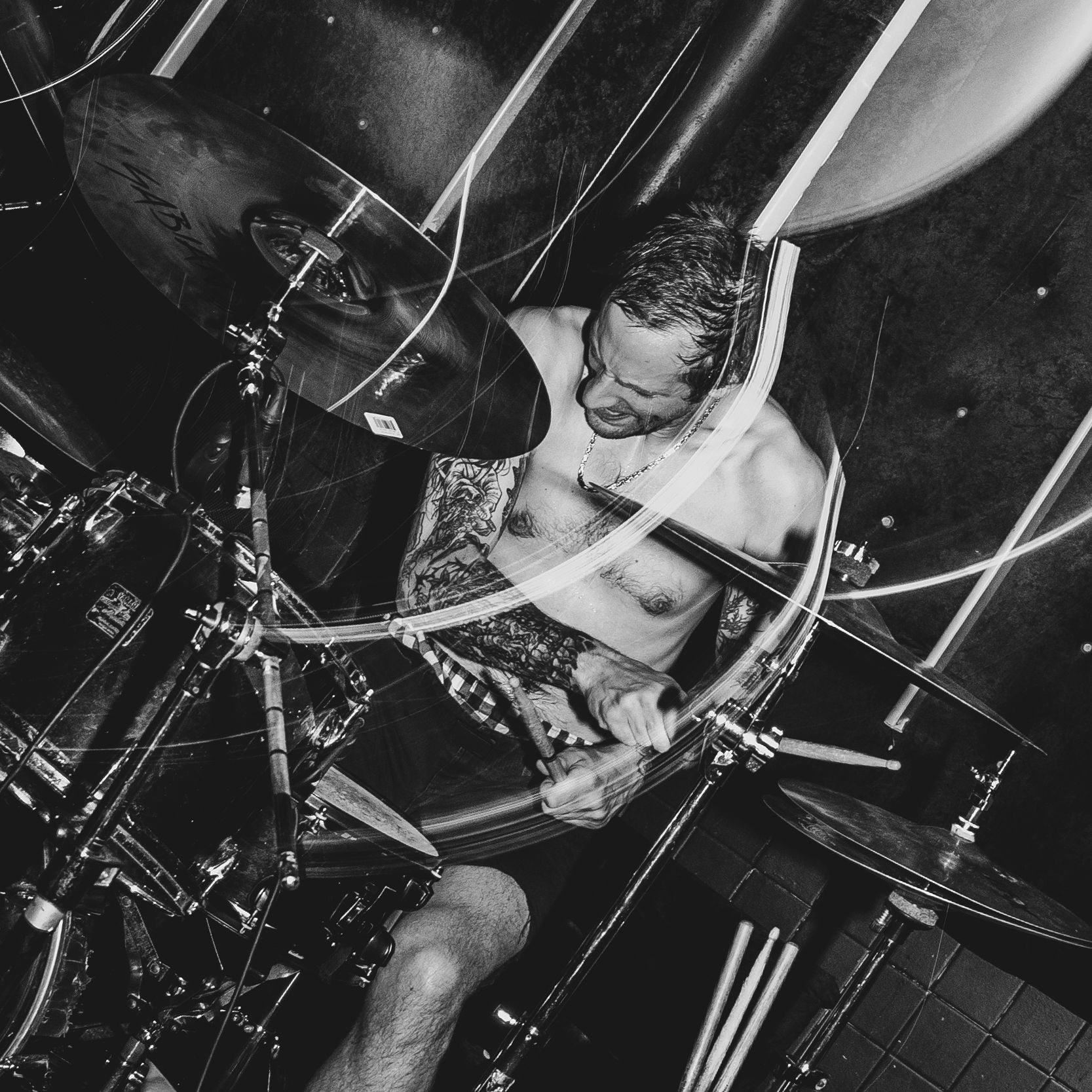 Black and white photo of a shirtless male drummer performing energetically behind a drum kit on stage.
