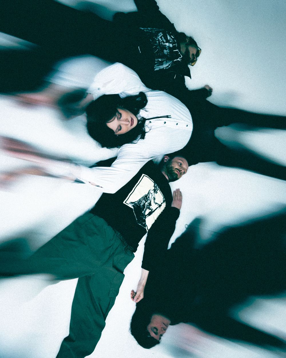 Four band members in dark clothing pose lying on a white backdrop, shot from above with a dramatic motion-blur effect.