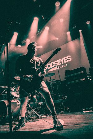 Guitarist performing on stage under teal lighting with Gods Eyes banner backdrop at concert.