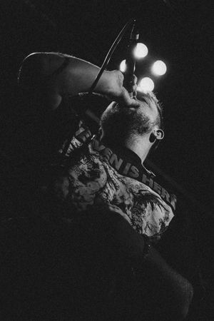 Black and white photo of a male vocalist screaming into a microphone under stage lights at a live concert.