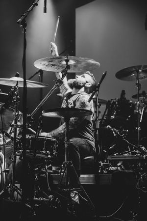 Black and white close-up of bearded drummer striking cymbal passionately during live performance.