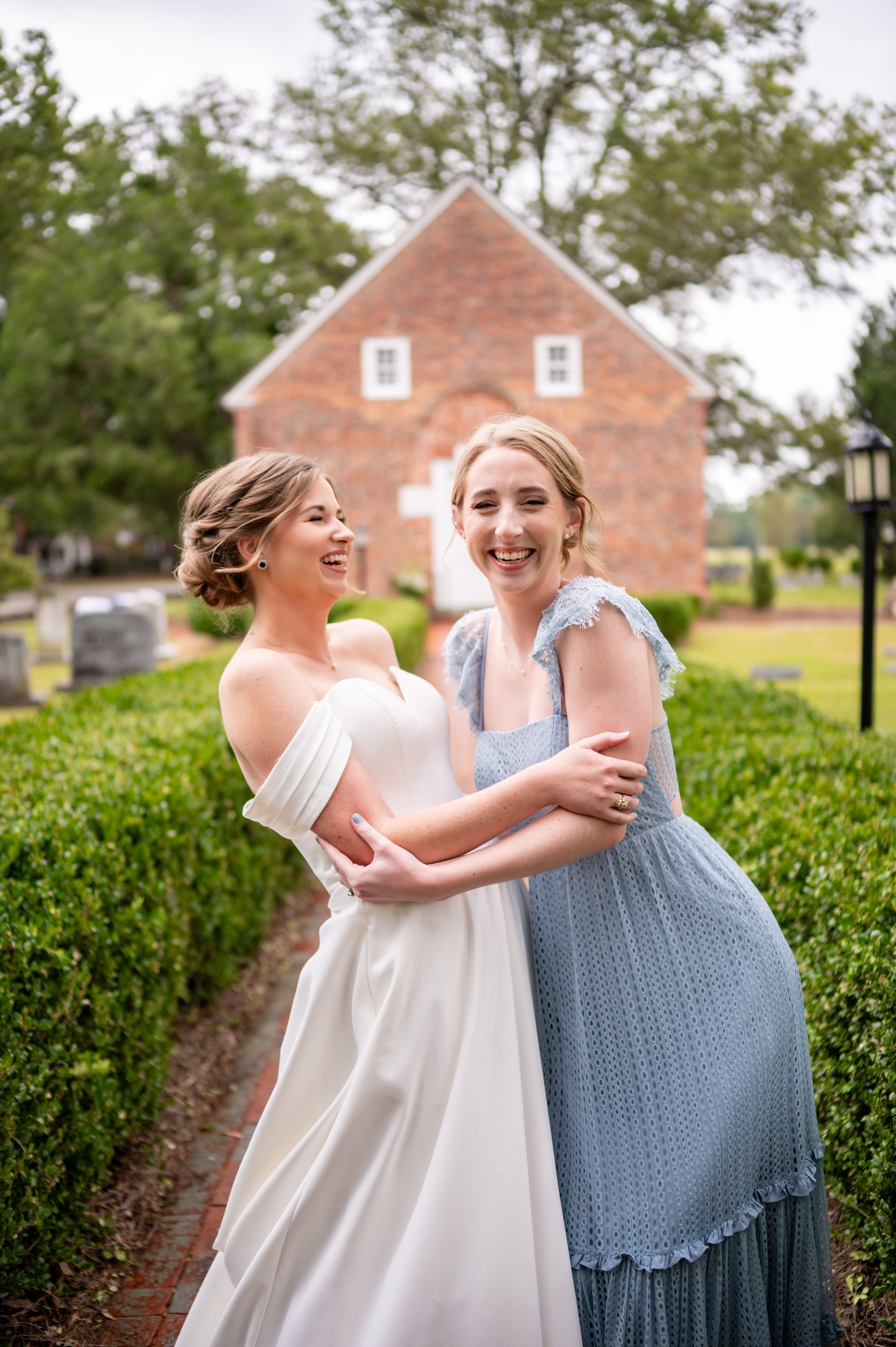 Two wedding attendants in white and blue dresses embrace joyfully in front of a brick building.