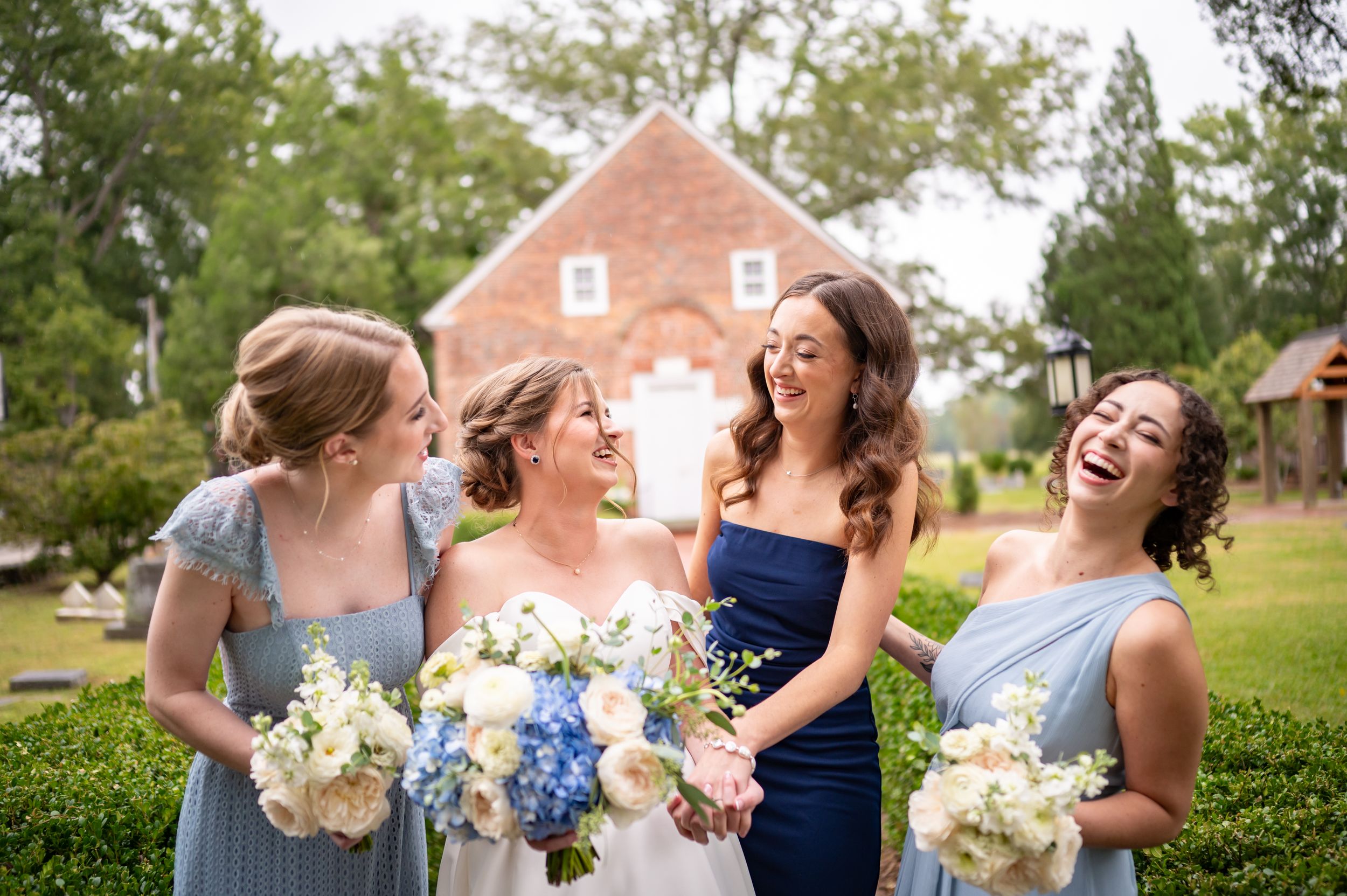 Wedding party with bridesmaids in soft blue dresses hold bouquets and share laughter outdoors.