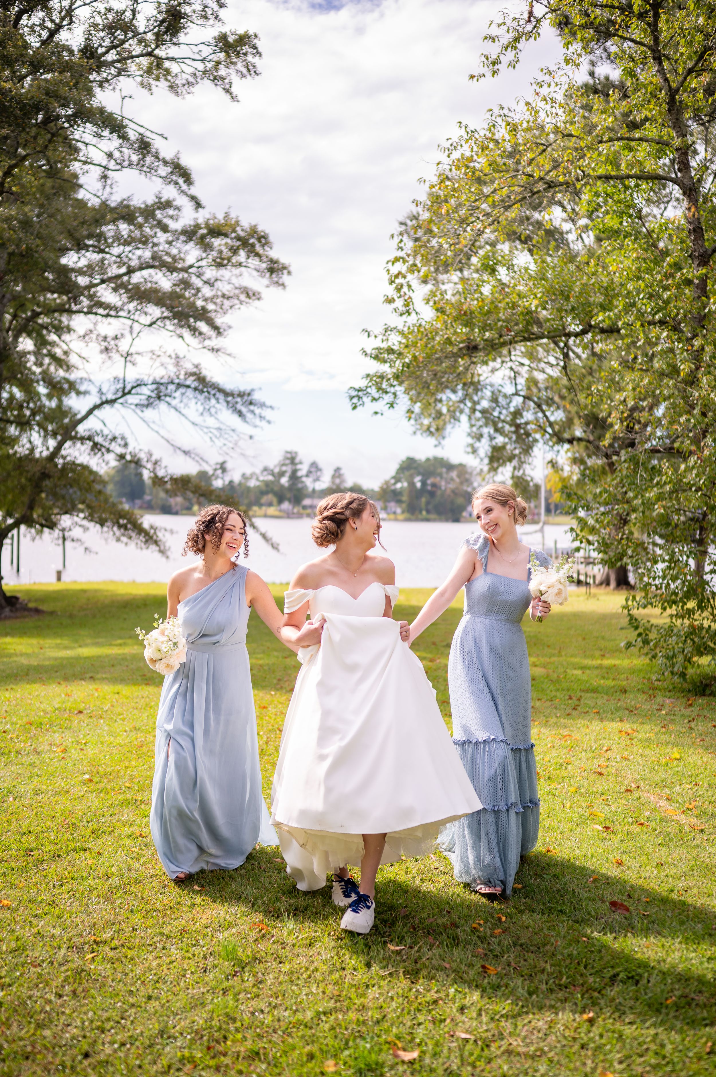 Three friends in flowing dresses dance together on a sunny lawn by the water.