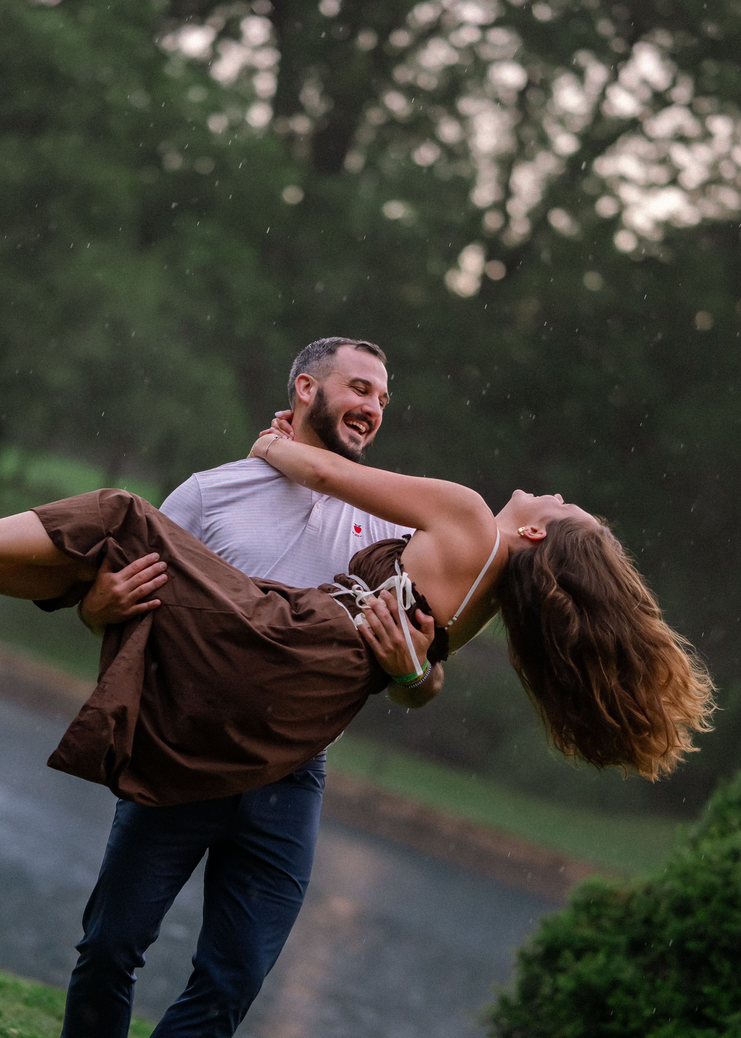 A joyful couple dancing in the rain, man lifting woman who laughs with hair flowing, in a lush green park.