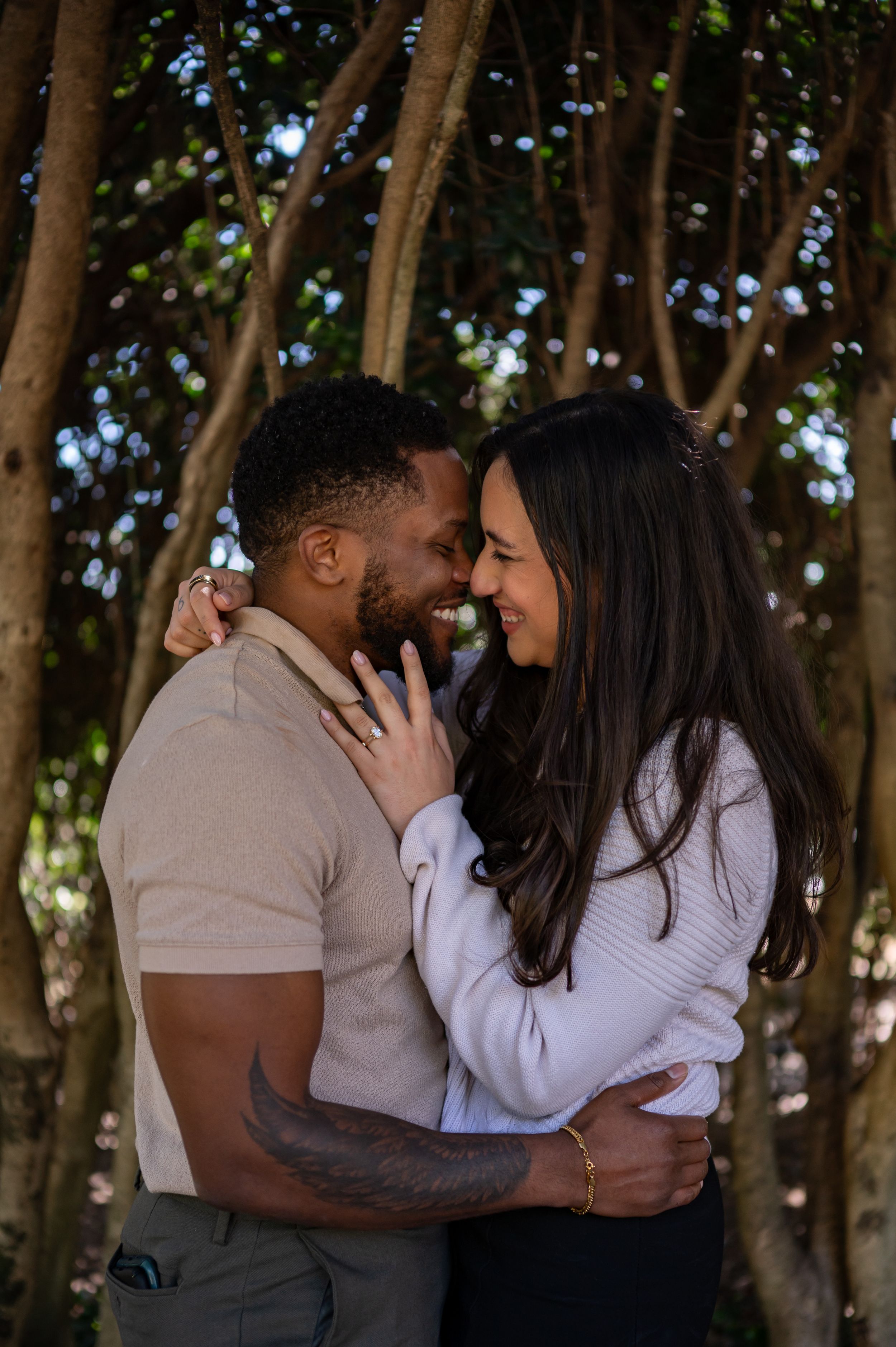 A joyful couple embraces and laughs together under a lush tree canopy.
