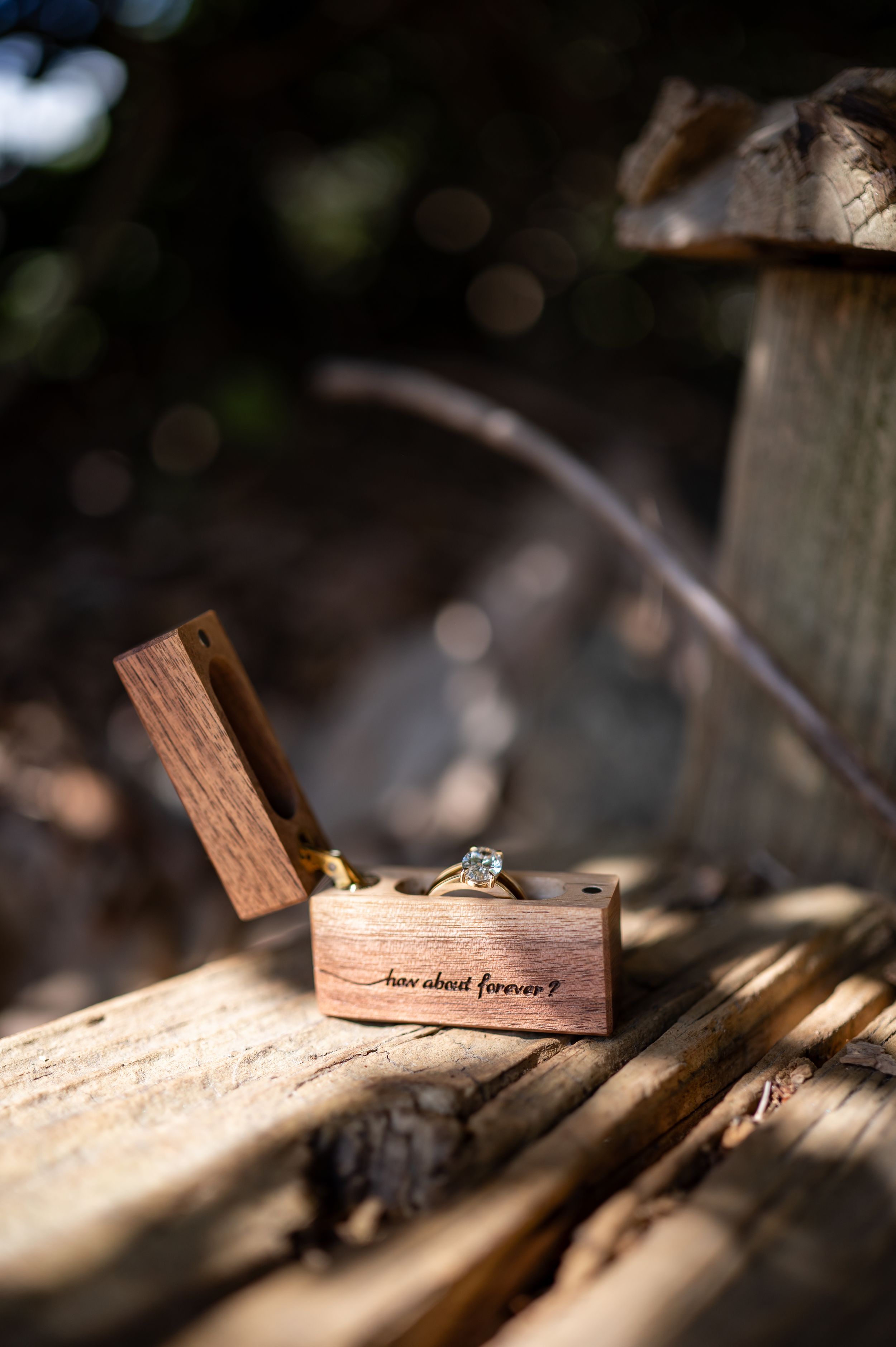 An engagement ring sits in a wooden box with engraved text, captured in natural sunlight.