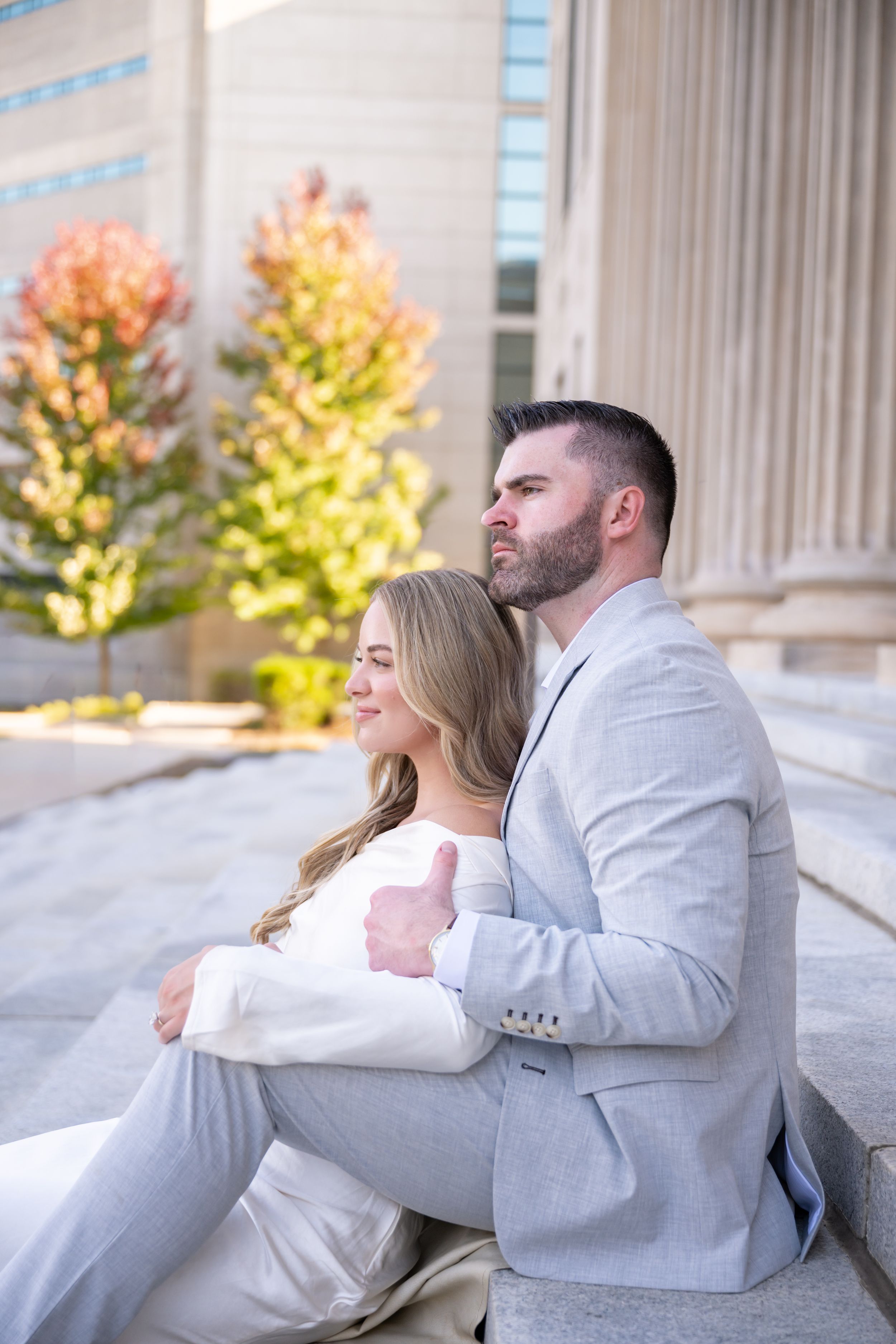 A couple in white and light gray attire share a playful moment on marble steps with autumn foliage in the background.