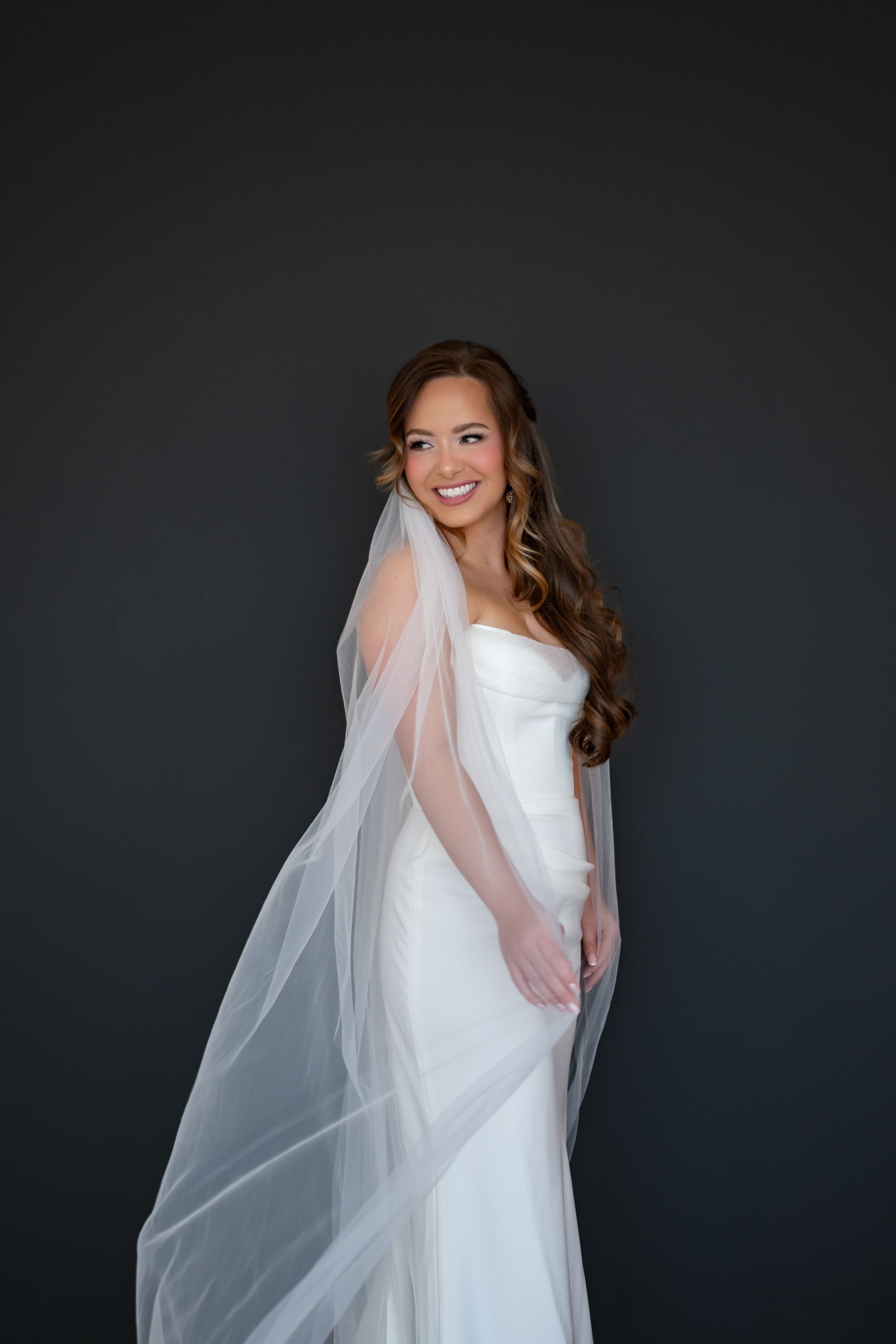 Bride in classic white mermaid wedding dress and long veil poses against dark background in elegant bridal portrait series.