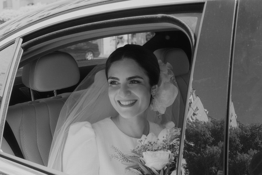 A black and white photo showing a bride smiling while seated in a car with flowers.