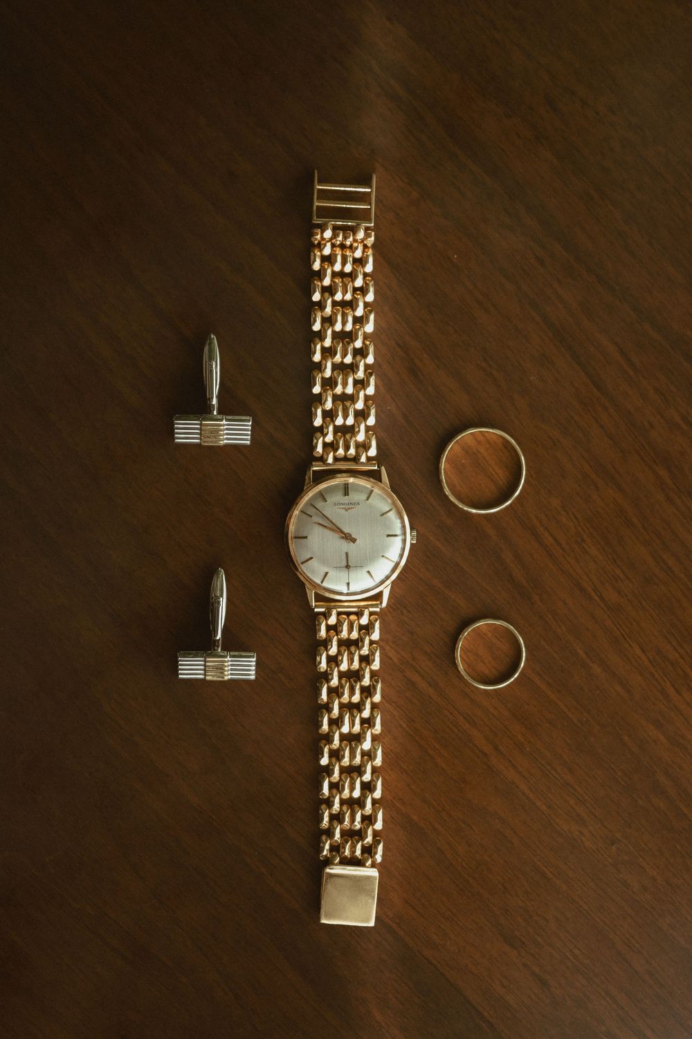 Gold wedding ring, cufflinks, and vintage watch with metal band arranged on dark wooden surface.