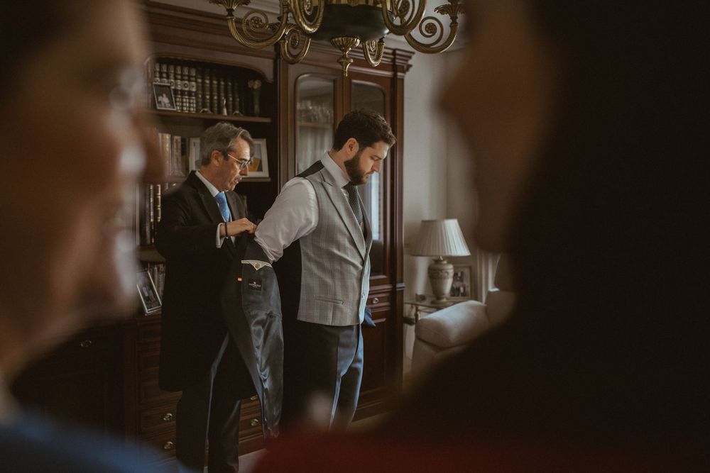 Two men in formal attire stand in an elegant wood-paneled room with vintage decor and chandelier lighting.