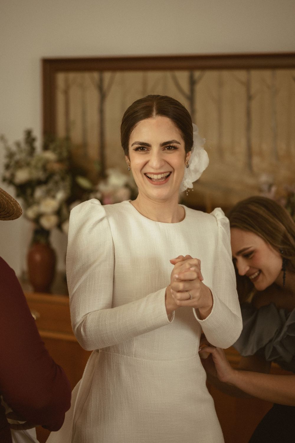 Elegant bride in a long-sleeved white wedding dress clasps her hands together with joy during her wedding celebration.