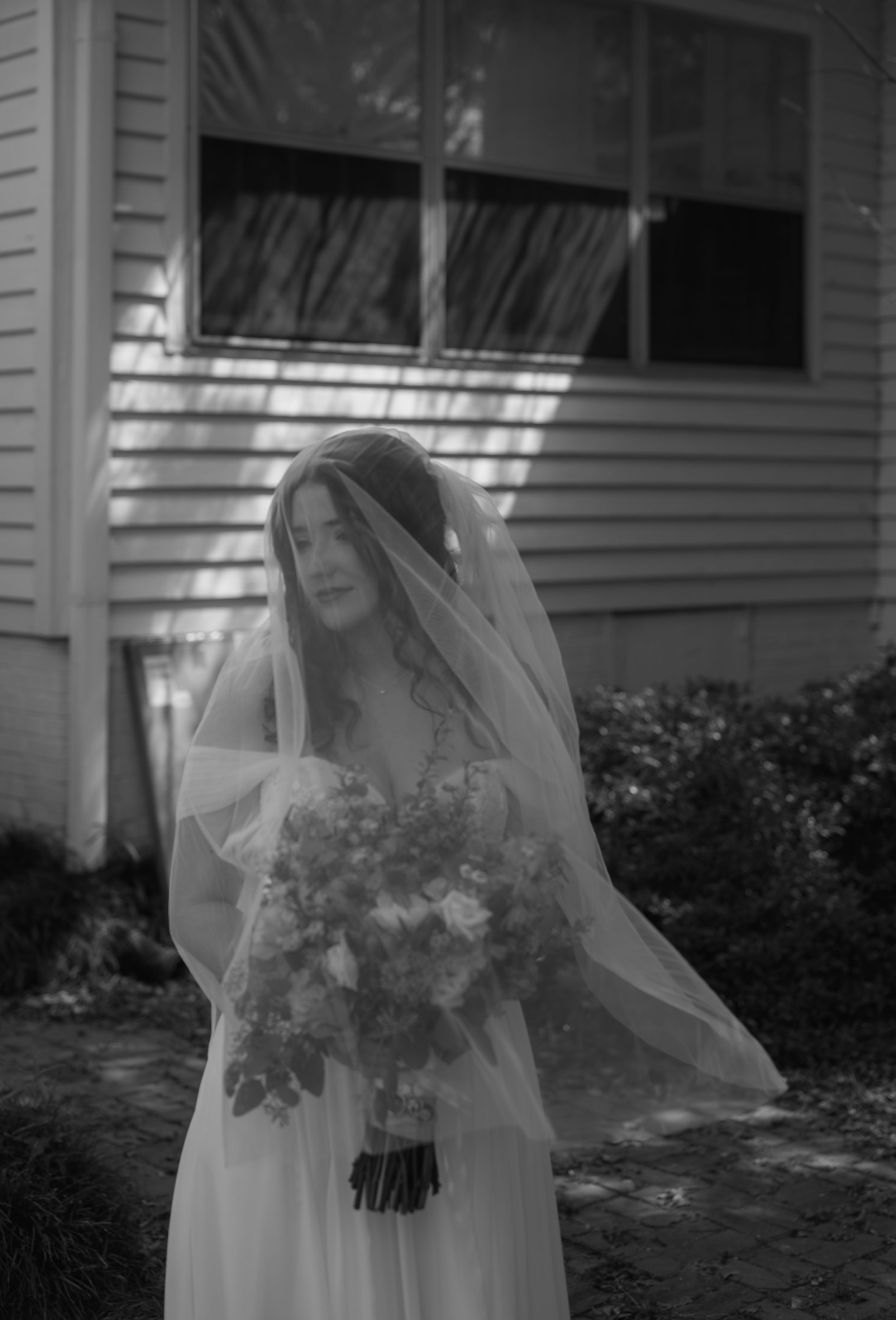 Ethereal black and white portrait of a bride holding a bouquet outside a white house.