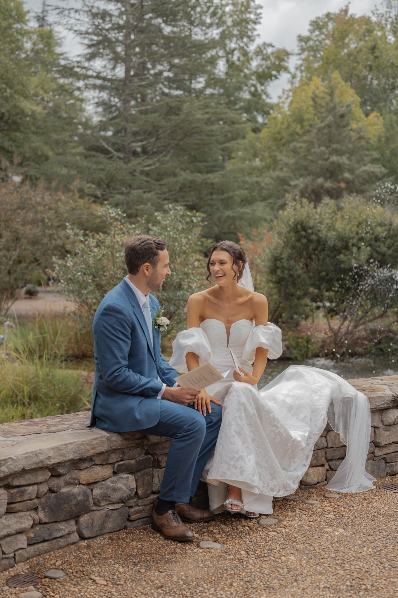 Couple shares intimate moment while sitting on stone wall surrounded by autumn trees.